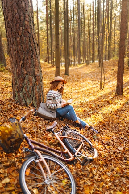 Una mujer elegante con un libro cerca de un árbol disfruta del clima otoñal en el parque al fondo de la bicicleta