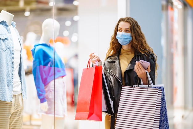 Mujer elegante en máscara médica protectora con bolsas de compras usando su teléfono Estilo de moda