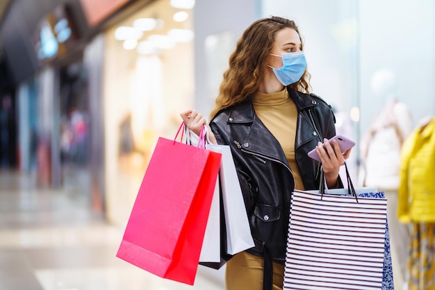 Mujer elegante con máscara médica protectora con bolsas de compras usando su teléfono Estilo de moda