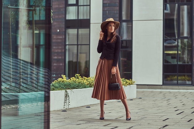 Mujer elegante de moda con chaqueta negra, sombrero marrón y falda con un bolso de mano caminando por el centro de una ciudad europea.