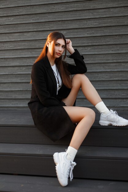 Mujer elegante modelo hermosa en un abrigo de moda negro con zapatillas blancas se sienta en las escaleras cerca de una casa de madera