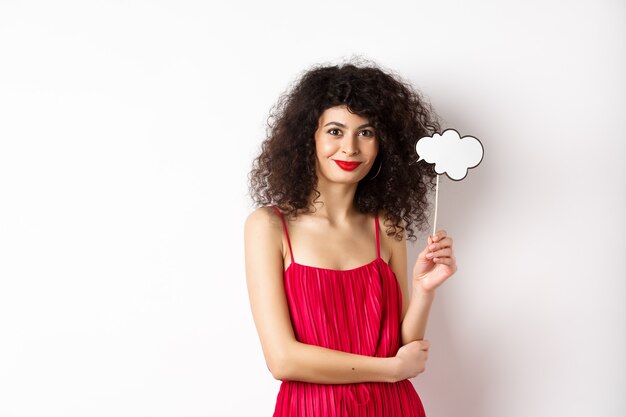 Mujer elegante con el pelo rizado, vestido de noche rojo, sosteniendo la nube y sonriendo, de pie sobre fondo blanco.