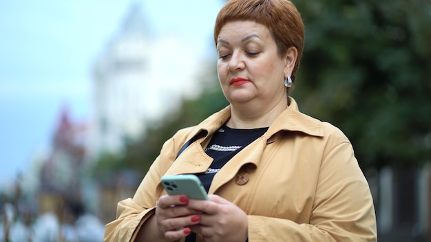 Una mujer elegante con el pelo rojo corto y un impermeable está parada en la calle y usando un teléfono inteligente