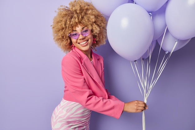 Mujer elegante positiva se prepara para la fiesta de las gallinas vestida con ropa elegante sostiene un montón de globos inflados de buen humor aislado sobre fondo púrpura Concepto de gente y celebración