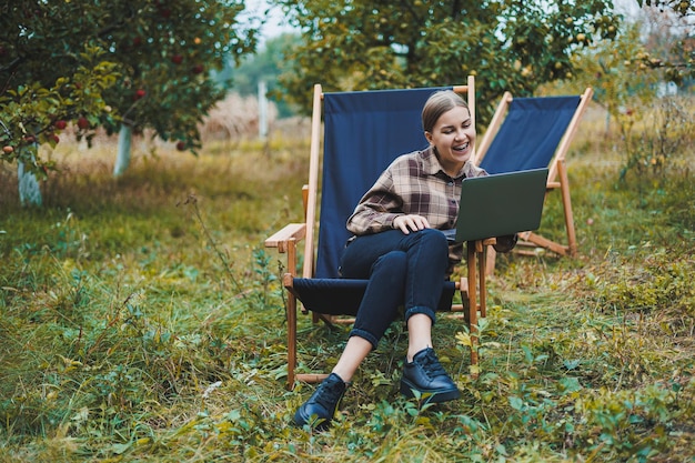 Mujer elegante sentada en una silla cerca de plantas verdes en el jardín y navegando por Internet en una computadora portátil mientras mira a un lado Mujer independiente de trabajo remoto