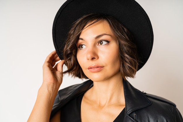 Mujer elegante con sombrero negro, vestido de noche y chaqueta de cuero posando en blanco.