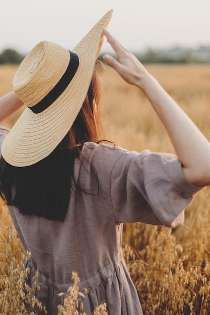 Mujer elegante con sombrero de paja de pie en el campo de avena en la vista posterior de la luz del atardecer Vida rural lenta