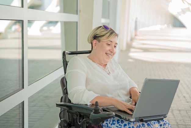 Una mujer elegante y sonriente con discapacidad sentada en una silla de ruedas disfrutando del trabajo de la computadora en línea