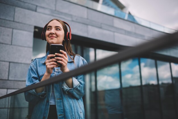 Mujer elegante sonriente positiva que disfruta de una nueva lista de reproducción con auriculares usando la aplicación en el teléfono inteligente