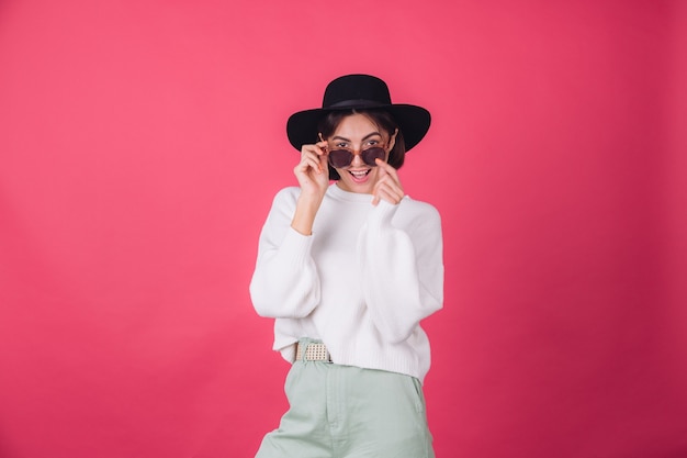 Mujer elegante en suéter blanco casual, gafas de sol y sombrero en la pared rosa roja