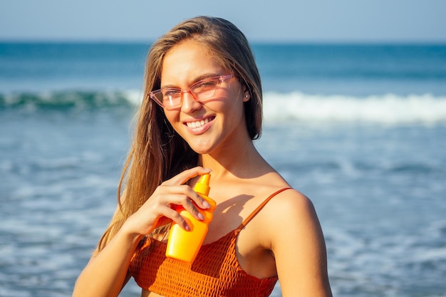 Mujer en un elegante traje de baño de bikini naranja coral aplicando protección solar en brazos y hombros bronceados sosteniendo una botella de spray solar spf contra el mar azul en las costas del Océano Índico