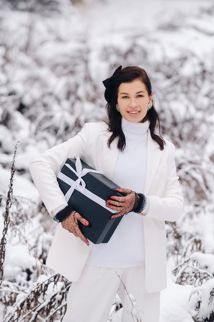 Una mujer elegante con un traje blanco con un regalo de Año Nuevo en sus manos en un bosque de invierno Una niña en la naturaleza en un bosque nevado con una caja de regalo