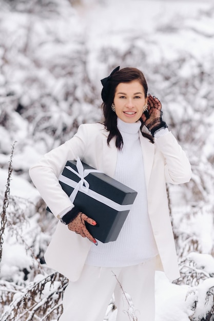 Una mujer elegante con un traje blanco con un regalo de Año Nuevo en sus manos en un bosque de invierno Una niña en la naturaleza en un bosque nevado con una caja de regalo