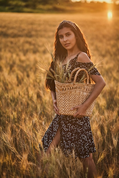 Mujer elegante en un vestido con cesta en el campo de trigo al atardecer.