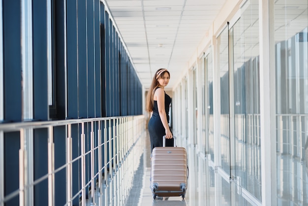 Mujer elegante con un vestido negro con una maleta en la terminal del aeropuerto esperando un avión