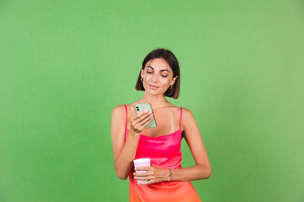 Mujer elegante en vestido de seda rosa aislado en verde feliz con una mirada de sonrisa en la pantalla del teléfono móvil, leer noticias del mensaje