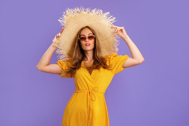 Mujer elegante en un vestido de verano amarillo claro gafas de sol y sombrero de paja posando en estudio de terciopelo púrpura ...