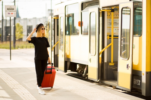 Mujer elegante visten total black con maleta de viaje roja se encuentra en la parada de la ciudad esperando el tranvía