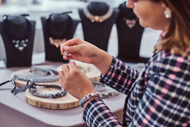 Mujer elegantemente vestida hace collares hechos a mano, trabajando con agujas e hilo en taller de joyería.