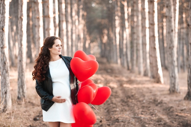 Mujer embarazada de 30 a 35 años sosteniendo globos rojos en forma de corazón de pie en el bosque