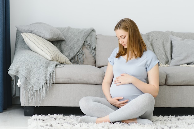 Mujer embarazada acariciando su vientre en casa. Joven rubia expectante sintiendo a su bebé empujar, sentada en el suelo, copie el espacio. Embarazo, descanso, vida, concepto de expectativa