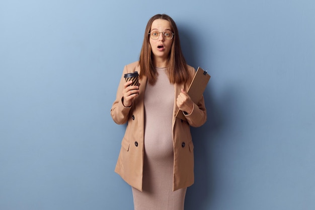 Foto mujer embarazada adulta joven con un vestido y una chaqueta posando aislada sobre un fondo azul sosteniendo papeles y café para llevar mirando a la cámara con la cara conmocionada