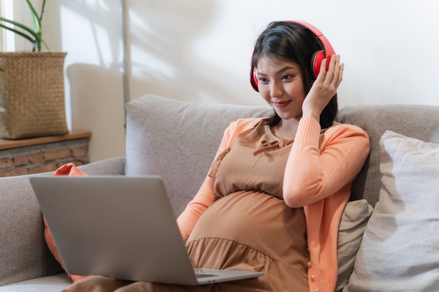 Una mujer embarazada de Asia sonríe y se sienta en el sofá y escucha música y usa la computadora portátil con una sensación feliz y relajada.