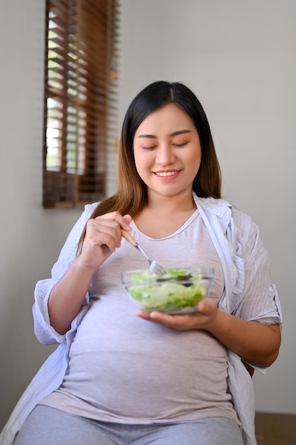 Mujer embarazada asiática feliz comiendo una ensalada saludable para el desayuno en una mesa junto a la ventana