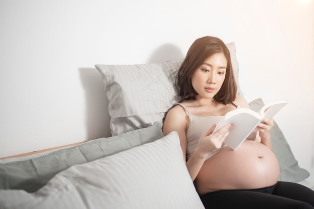 Mujer embarazada asiática hermosa que miente en su cama y que lee un libro