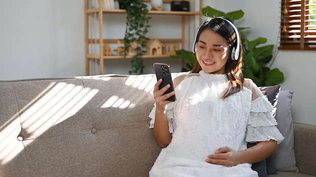 Mujer embarazada asiática relajada usando auriculares escuchando música en su sala de estar