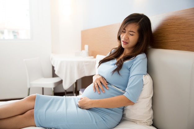 Mujer embarazada asiática tiene dolor de estómago sentado en su cama