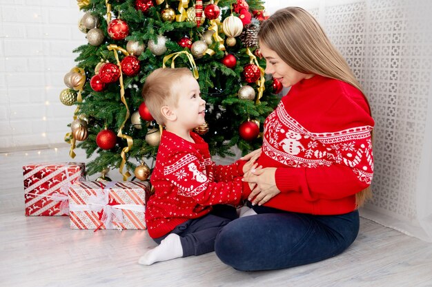 Una mujer embarazada con un bebé con un suéter rojo bajo el árbol de Navidad en casa abrazándose felicitándose y disfrutando del año nuevo y la Navidad