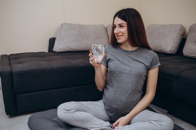 Mujer embarazada bebiendo agua sentada en el sofá Joven mujer embarazada tiene descanso en casa con espacio de copia de vidrio Concepto de sed de atención médica durante el embarazo