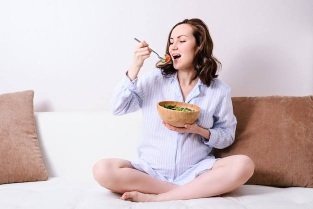 Foto mujer embarazada en camisa azul sentada con las piernas cruzadas en el sofá comiendo ensalada de verduras saludables en un tazón de madera. concepto de embarazo saludable