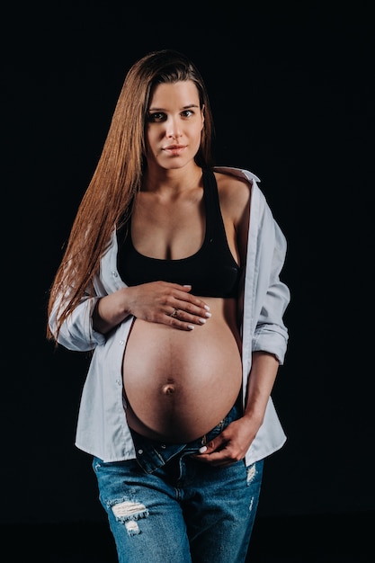 Mujer embarazada en camisa blanca y jeans en estudio sobre fondo negro.