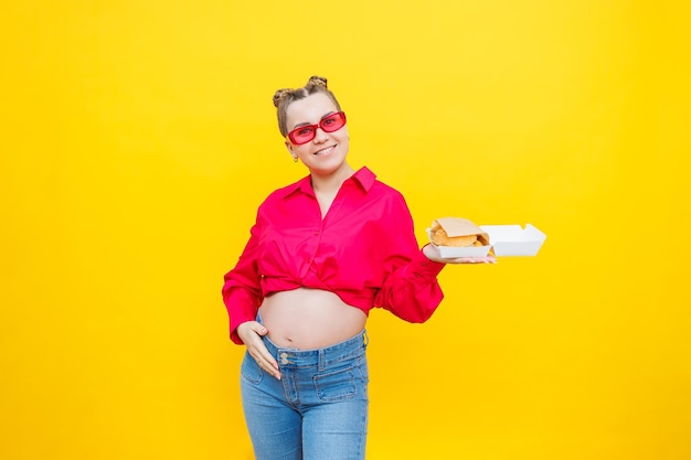 Una mujer embarazada con una camisa rosa y jeans sostiene una sabrosa hamburguesa Comida dañina durante el embarazo Concepto de embarazo fácil y feliz