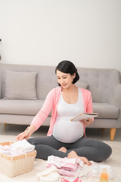 Una mujer embarazada en casa el piso revisando la lista ropa de bebé preparándose para ir al hospital de maternidad | Foto Premium