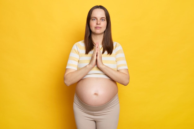 Mujer embarazada caucásica concentrada en ropa casual posando aislada sobre un fondo amarillo mirando a la cámara con un gesto de oración tratando de relajarse