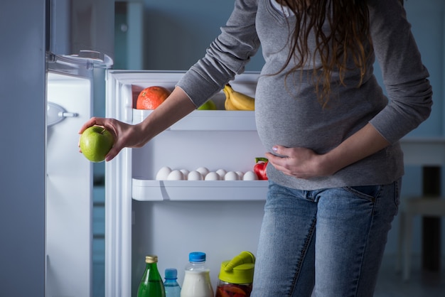 Mujer embarazada cerca de nevera buscando comida y aperitivos por la noche