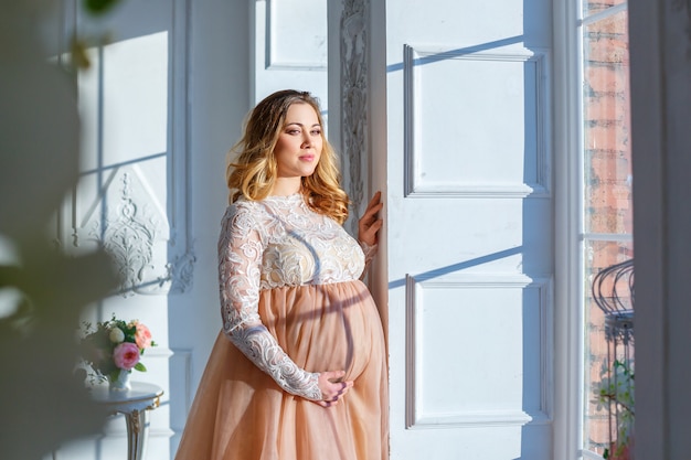 Una mujer embarazada cerca de la ventana con un hermoso vestido, maternidad.