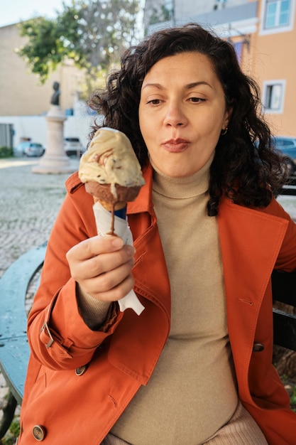 Mujer embarazada comiendo un cono de helado de chocolate afuera en un banco