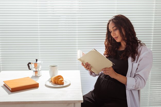 Mujer embarazada comiendo y leyendo un libro en casa. Concepto de mañana, desayuno y embarazo.