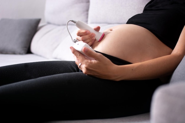 Mujer embarazada con un doppler fetal escuchando el corazón del bebé sentado en el sofá de casa