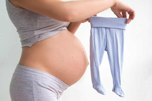 Mujer embarazada elige ropa para el bebé. La mujer embarazada sostiene en sus manos pantalones de niños pequeños. Deslizadores para niños pequeños. Ropa para el recién nacido. Esto es un chico. El embarazo. Maternidad.