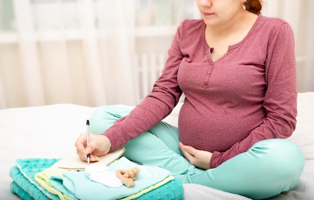 Una mujer embarazada escribe una lista y prepara ropa de bebé para el hospital de maternidad.