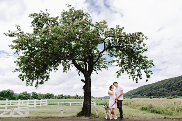 Mujer embarazada con esposo y perro husky cerca de un árbol en la naturaleza