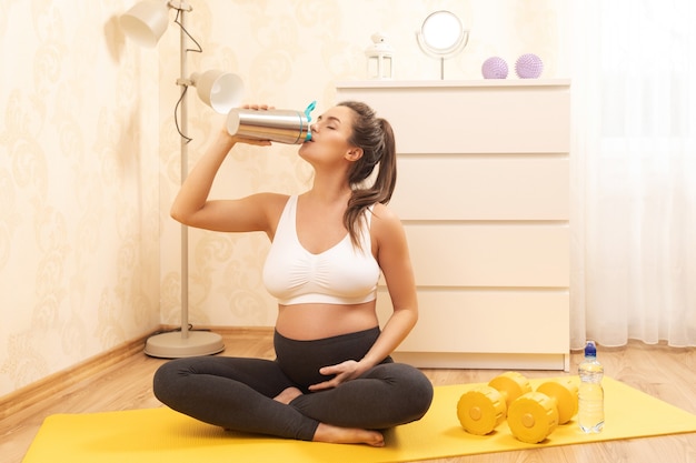 La mujer embarazada está bebiendo batido de proteínas después de su entrenamiento físico en casa