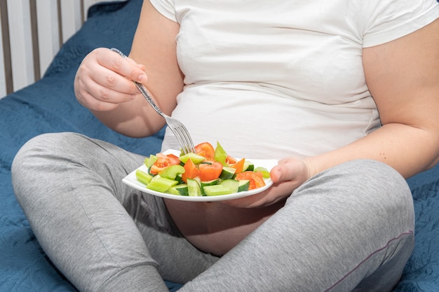 Una mujer embarazada está comiendo una ensalada de frutas orgánicas caseras saludables mientras está sentada en su cama. Concepto de nutrición y embarazo saludable