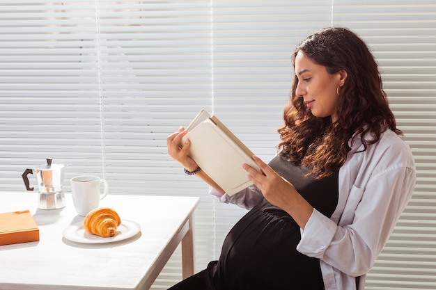 La mujer embarazada está leyendo el libro durante el desayuno. Concepto de embarazo y educación