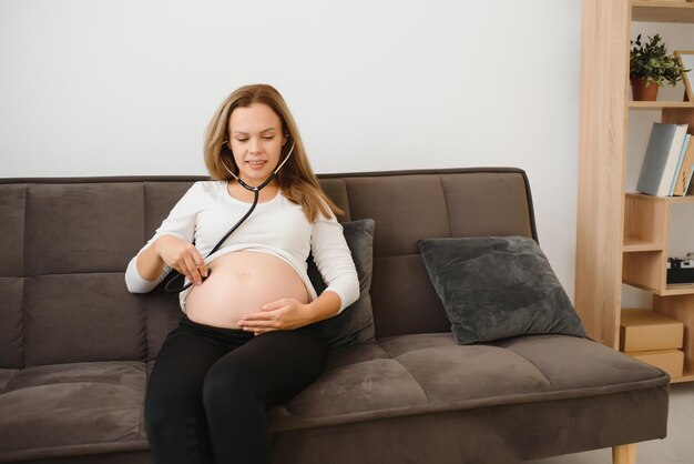 Mujer embarazada con estetoscopio examinando su vientre en el sofá, concepto de mujer embarazada feliz.